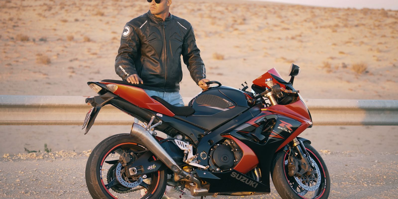 Man wearing a leather jacket and cap posing with a sports motorcycle on a desert road