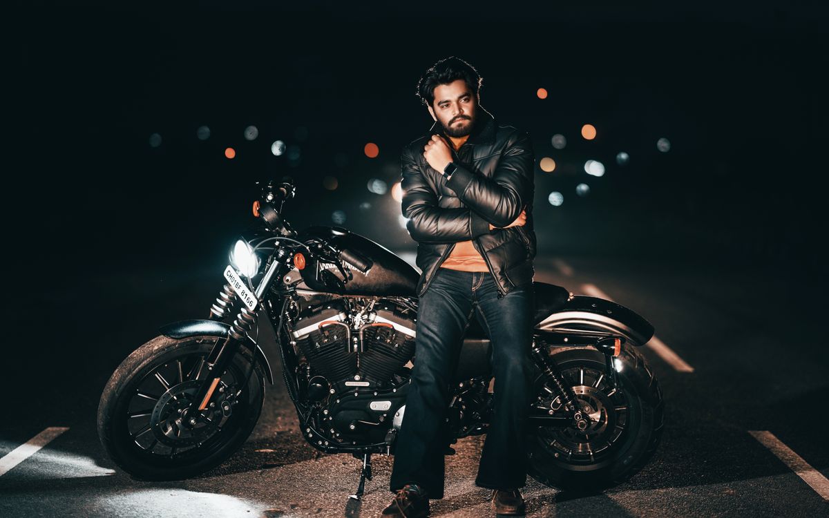 Man in a leather jacket posing with a motorcycle at night with city lights in the background