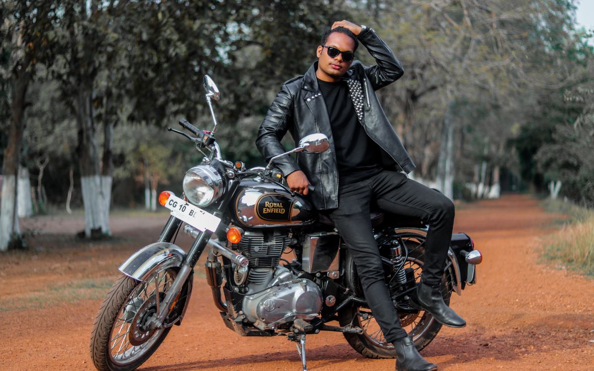 Young man posing with a motorcycle on a dirt road, showcasing a fitted leather jacket