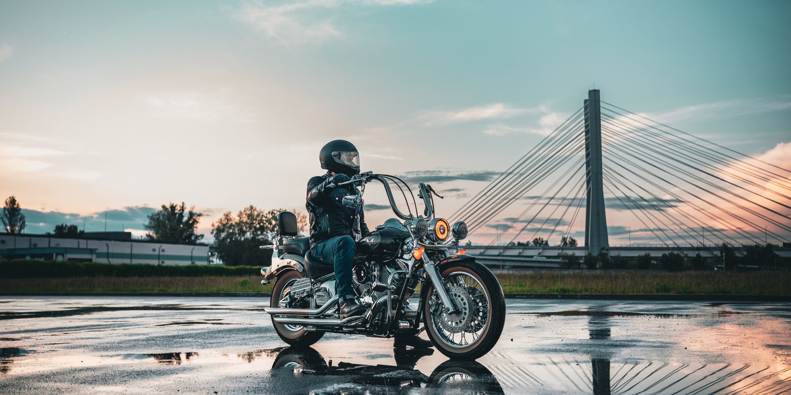 Motorcycle rider in leather gear on a wet road with a bridge in the background during sunset
