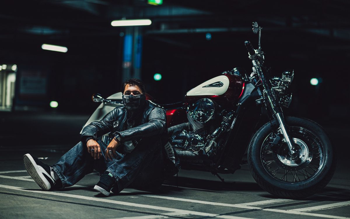 Biker in a leather jacket kneeling beside a motorcycle in a dimly lit garage