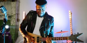Guitarist playing electric guitar on stage with vibrant lights, wearing a leather jacket and flat cap