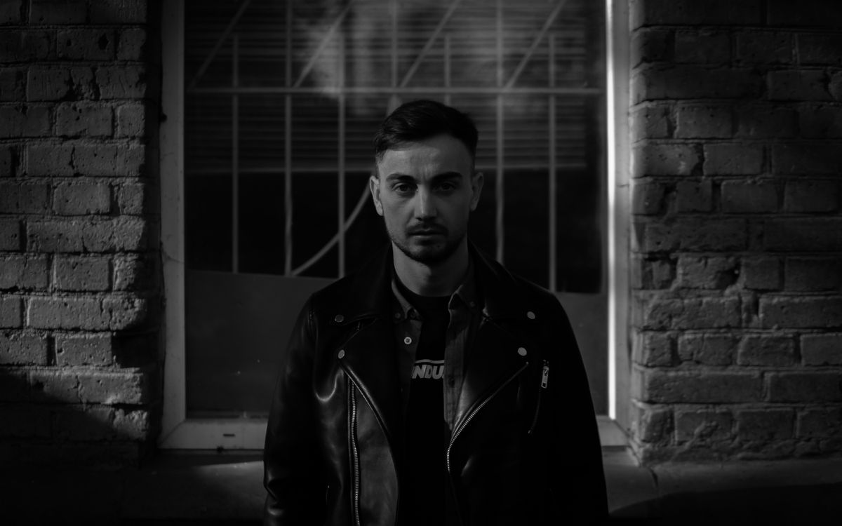 Monochrome portrait of an adult man in a leather jacket standing against a brick wall