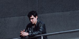 A man in a black leather jacket standing thoughtfully by a railing in an urban setting