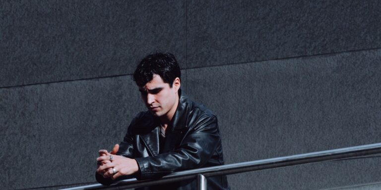 A man in a black leather jacket standing thoughtfully by a railing in an urban setting