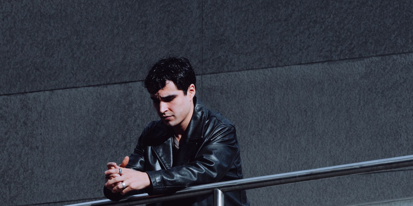A man in a black leather jacket standing thoughtfully by a railing in an urban setting