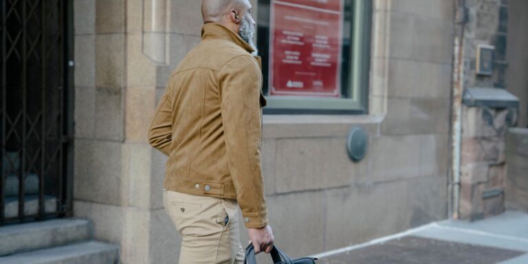 A man with a beard walks down a city street carrying a briefcase, wearing a beige jacket