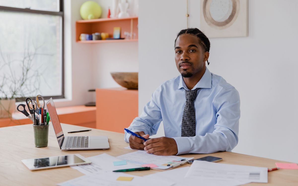 A professional businessman with a beard and braided hair sits at his desk with a laptop and documents, working diligently