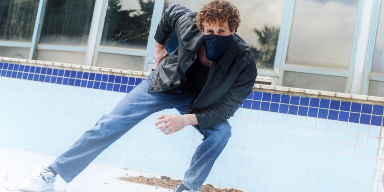 Man in a leather jacket and jeans leaning against a wall with a stylish, relaxed pose