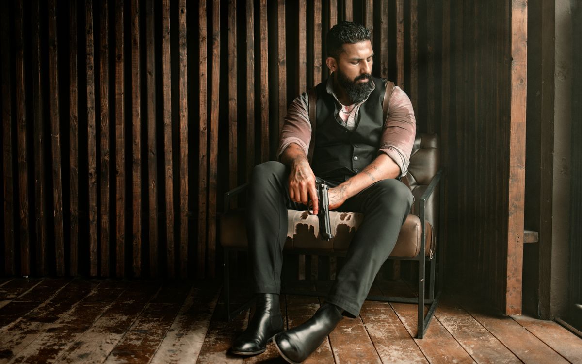 Bearded man in a vest seated indoors against a rustic backdrop