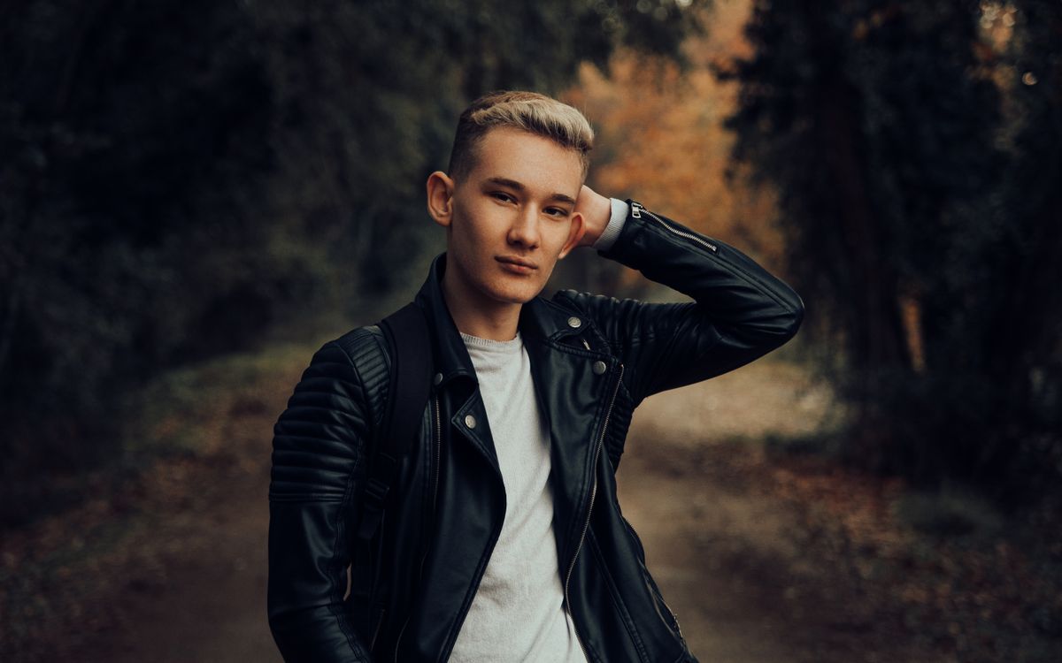 A young man in a leather jacket poses in a forest setting during autumn
