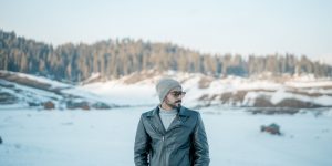 A man in a leather jacket standing among snow covered mountains under a clear sky