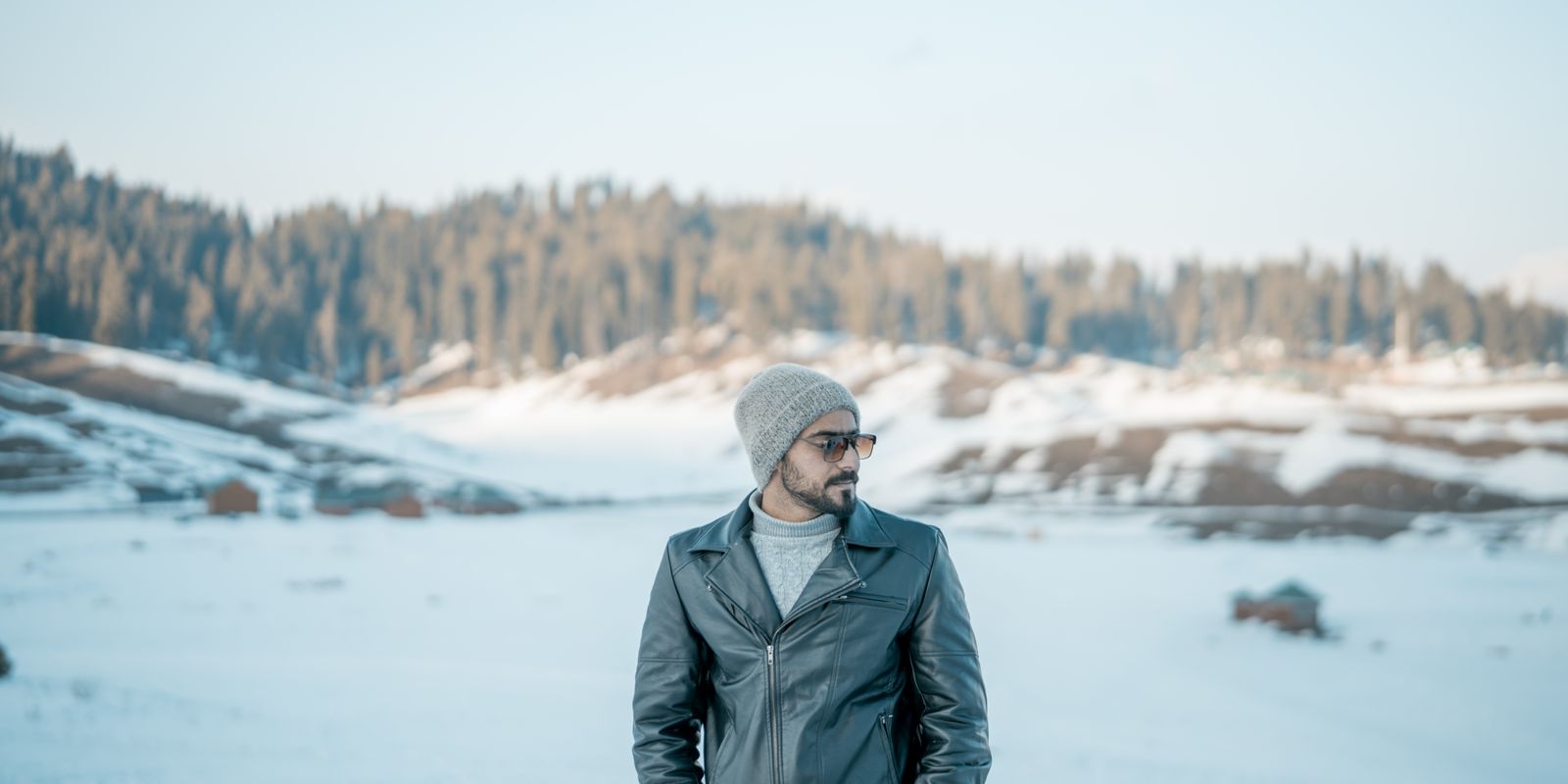 A man in a leather jacket standing among snow covered mountains under a clear sky