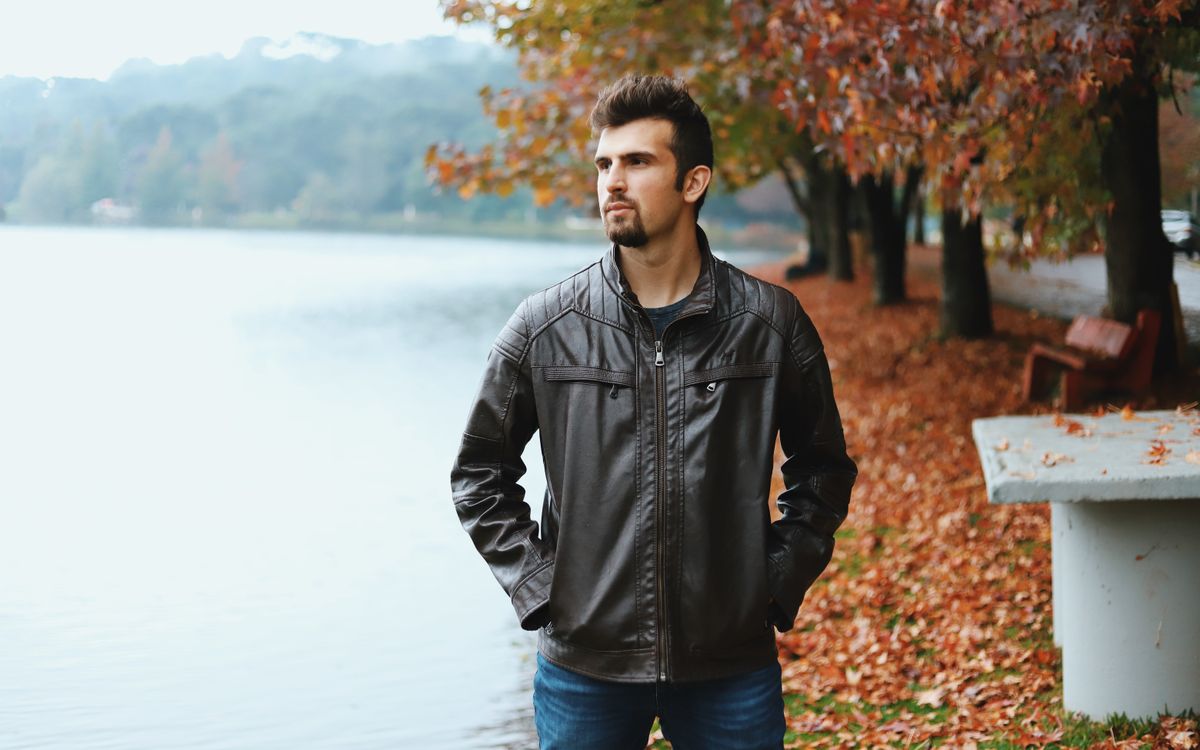 A bearded man in a brown leather jacket standing by a lake during autumn
