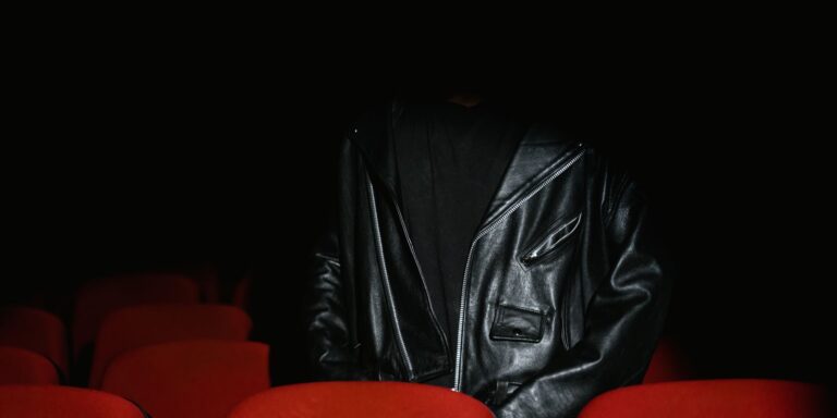 A person in a black leather jacket seated in a dimly lit cinema with red seats