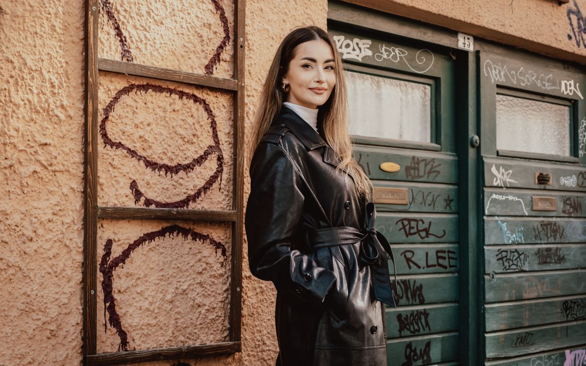 A woman posing confidently in a long black leather coat against an urban wall