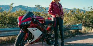 Woman in a red leather jacket standing beside a red motorcycle on a mountain road