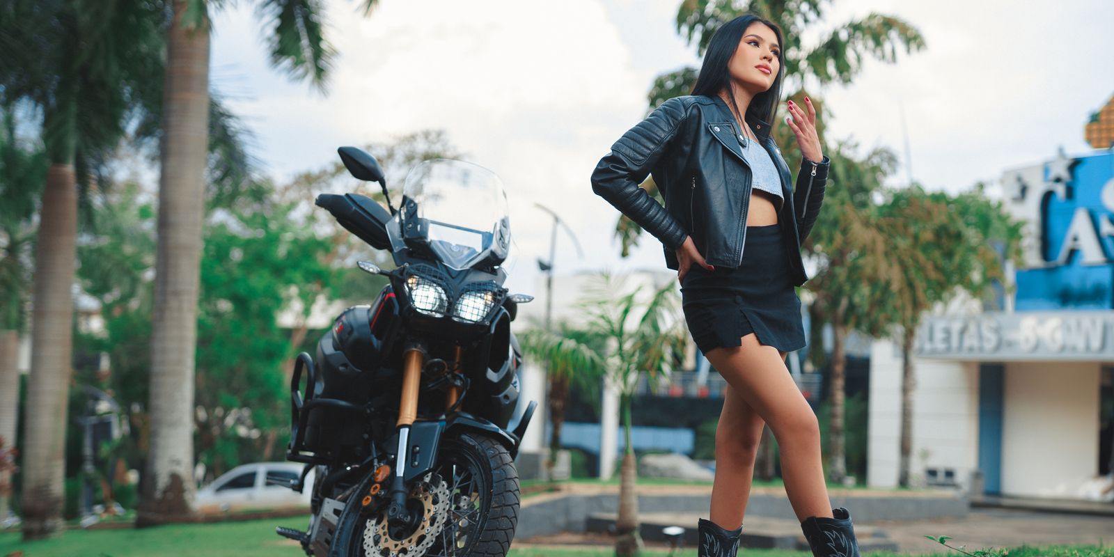 Confident woman in a leather biker jacket standing beside a motorcycle in the sunshine