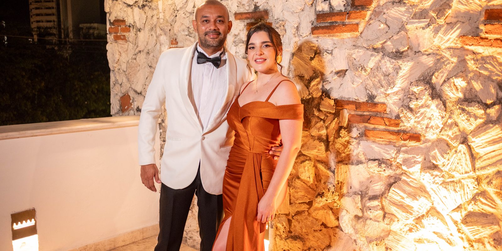 Stylish couple in formal evening attire posing together against an illuminated stone wall