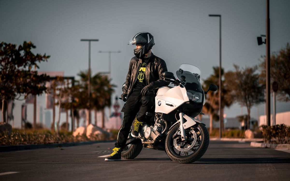 A rider in black gear stands beside a motorcycle in daylight