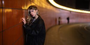 A woman in a leather jacket poses against an illuminated tunnel wall, a moody urban look