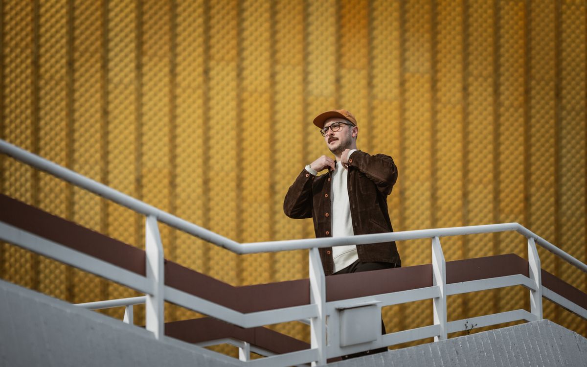 A fashionable man in urban attire stands on a contemporary staircase with a textured wall