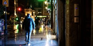 A man walks along a wet, neon lit city street in the rain at night