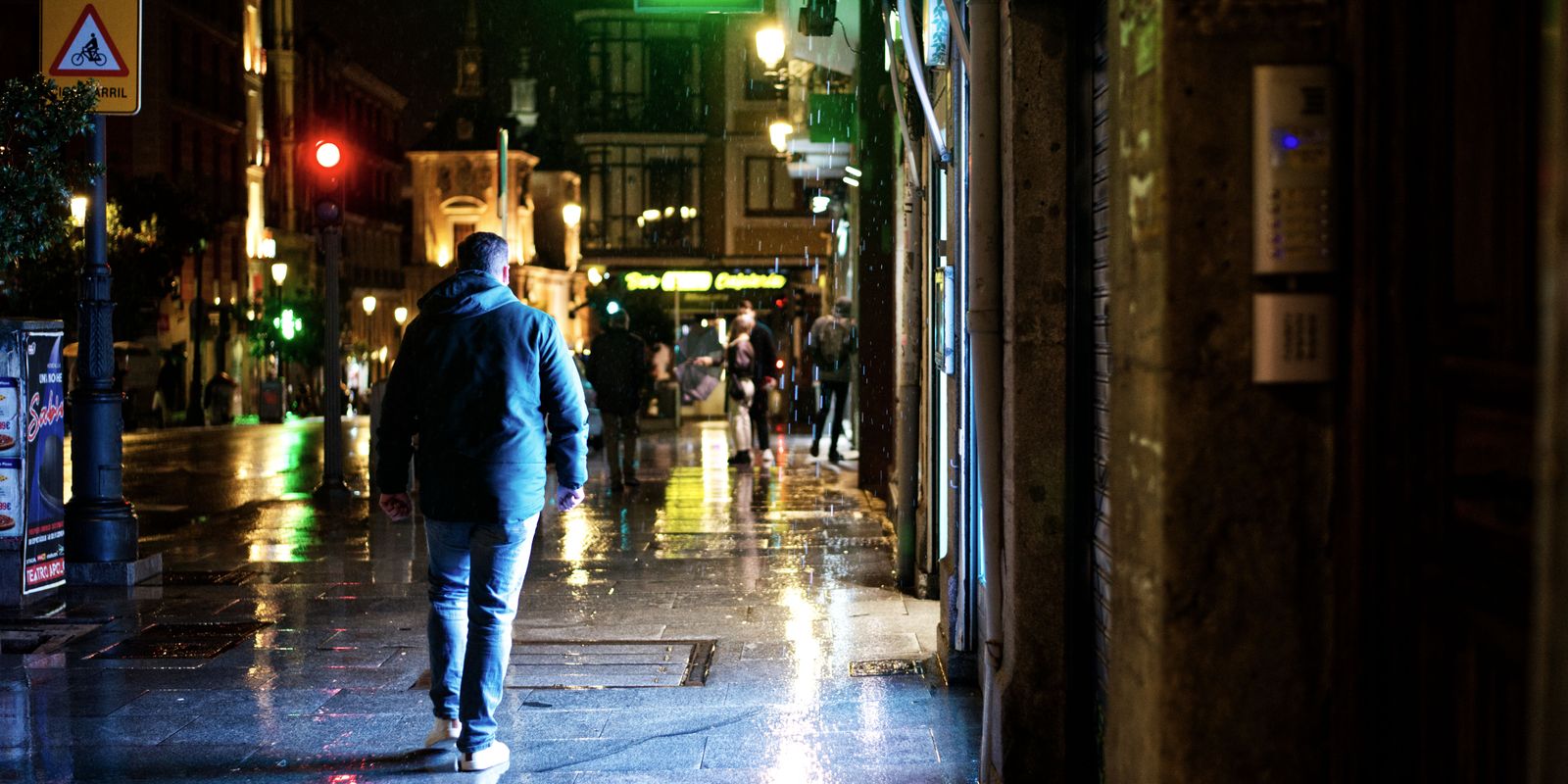 A man walks along a wet, neon lit city street in the rain at night