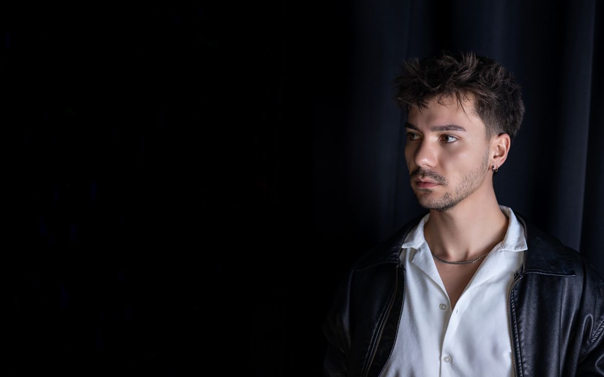 Man in a leather jacket posing indoors against a black curtain