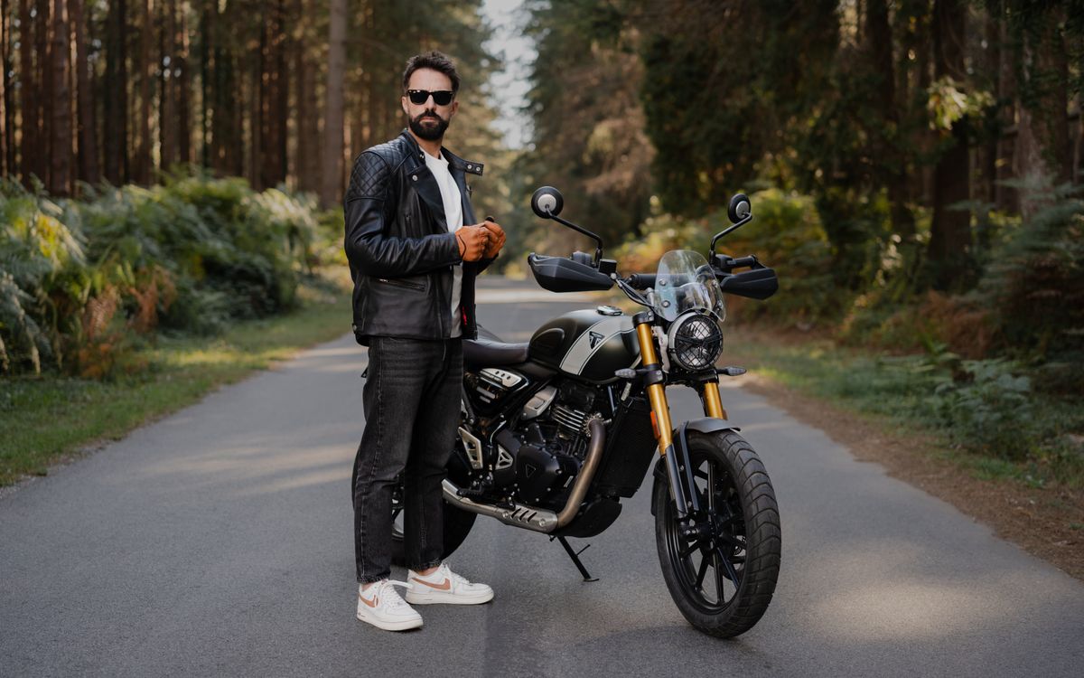A man in a leather jacket standing beside a motorcycle on a quiet forest road