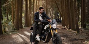 A man in a brown leather jacket pausing with a motorcycle on a quiet forest trail