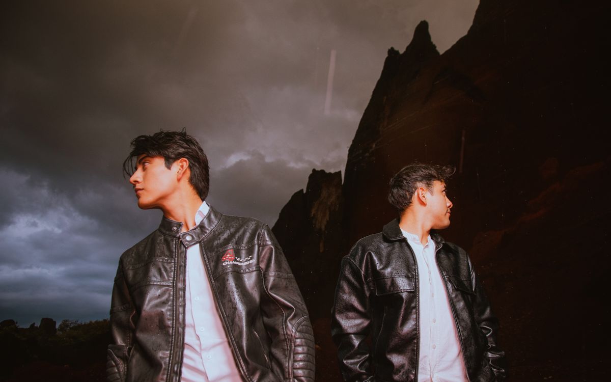 Two men in brown leather jackets outdoors under a dramatic, moody sky