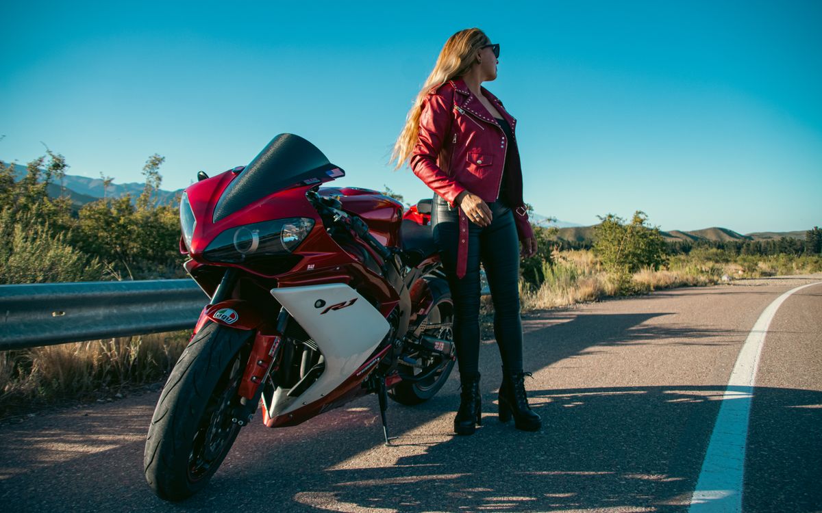 A woman in a red leather jacket standing beside a red motorcycle on an open road
