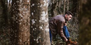 A man in a brown leather jacket tying brown leather boots on a wooded path