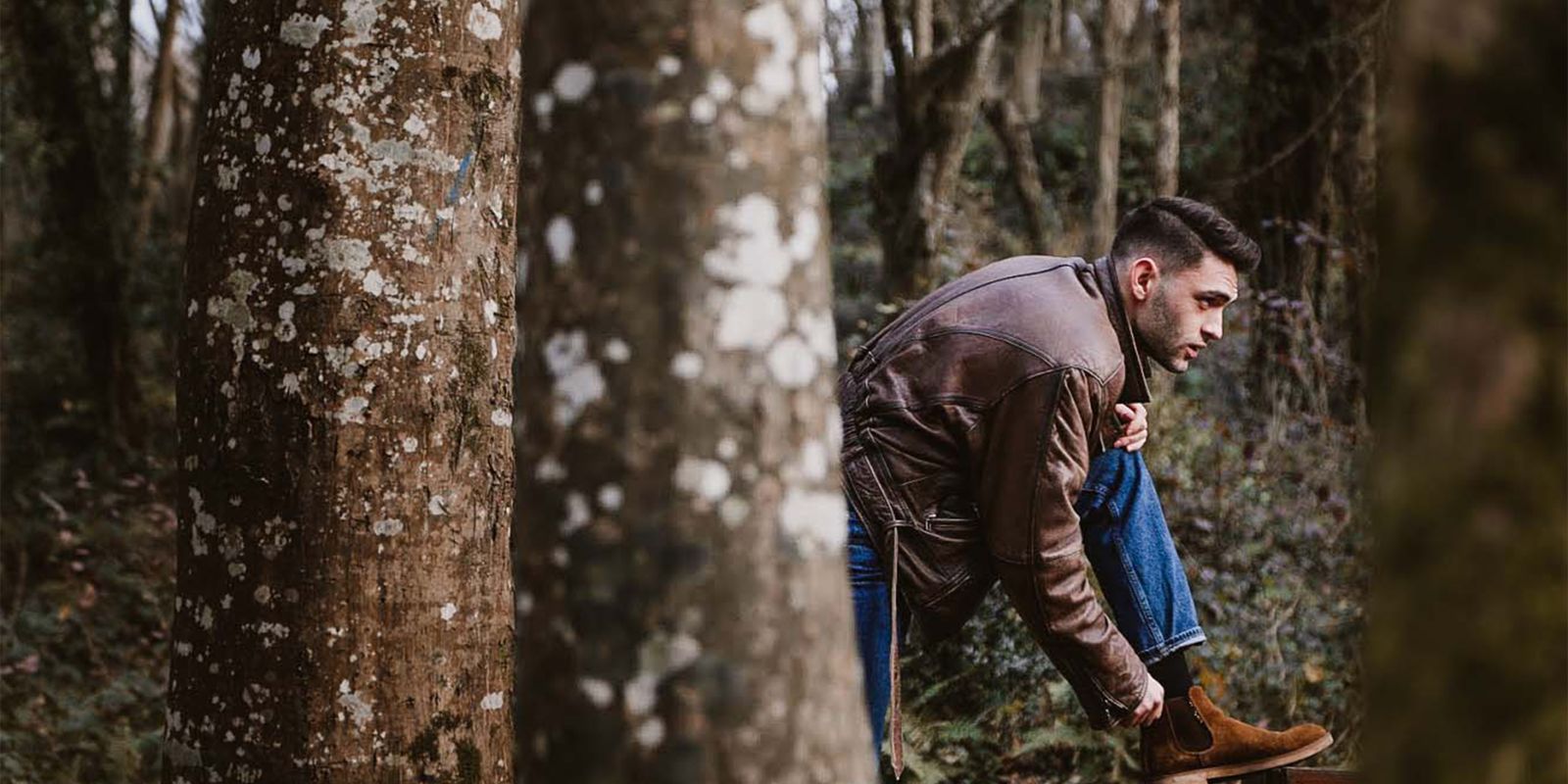A man in a brown leather jacket tying brown leather boots on a wooded path